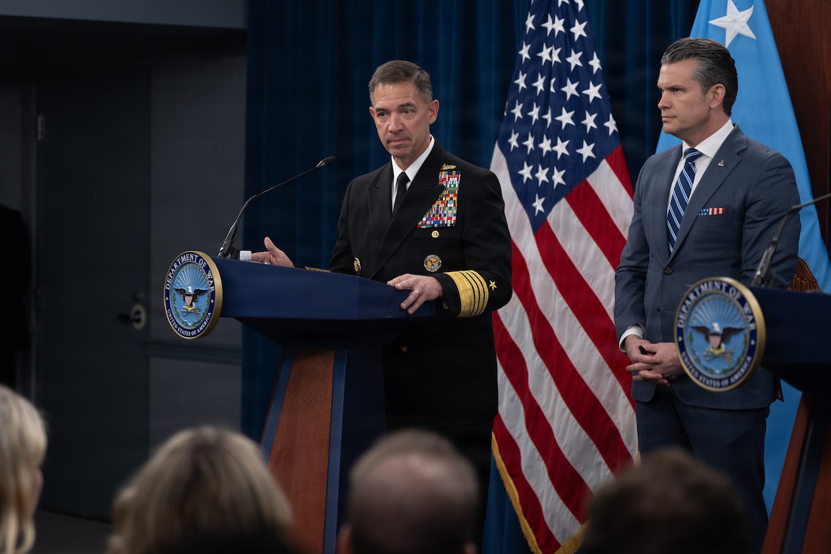 A man in a military uniform stands behind a lectern. A man in civilian attire stands near him. Behind them both are two flags.