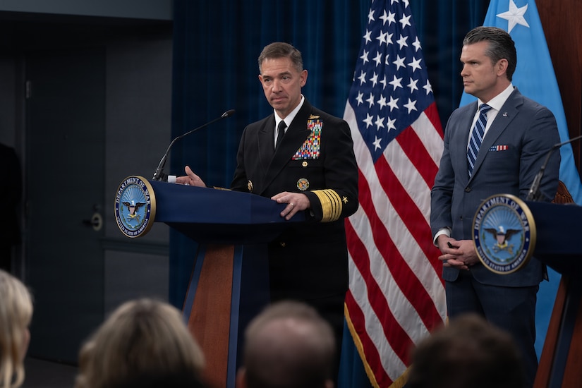 A man in a military uniform stands behind a lectern. A man in civilian attire stands near him. Behind them both are two flags.