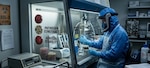 A laboratory technician wearing full protective gear works inside a biosafety cabinet in a high-containment lab. The worker handles glass equipment and samples while surrounded by petri dishes and scientific instruments. Signs on the cabinet reference “Pathogen Construct” and “Aerosol Stability,” and a “BSL-3 Access” warning is visible, indicating research involving potentially hazardous biological materials.