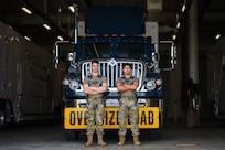 Two airmen stand in front of a payload transporter replacement.