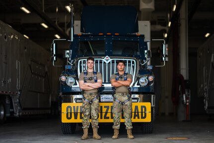 Two airmen stand in front of a payload transporter replacement.