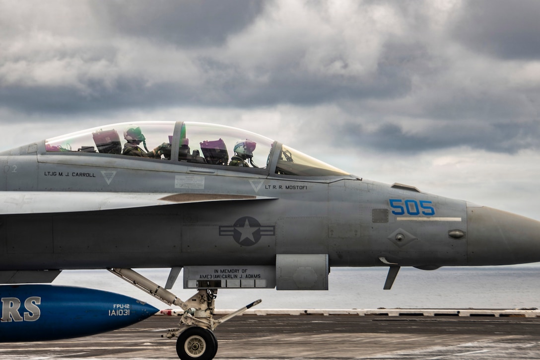 A side view of sailors wearing helmets and masks in an aircraft aboard a ship at sea under a gloomy sky.