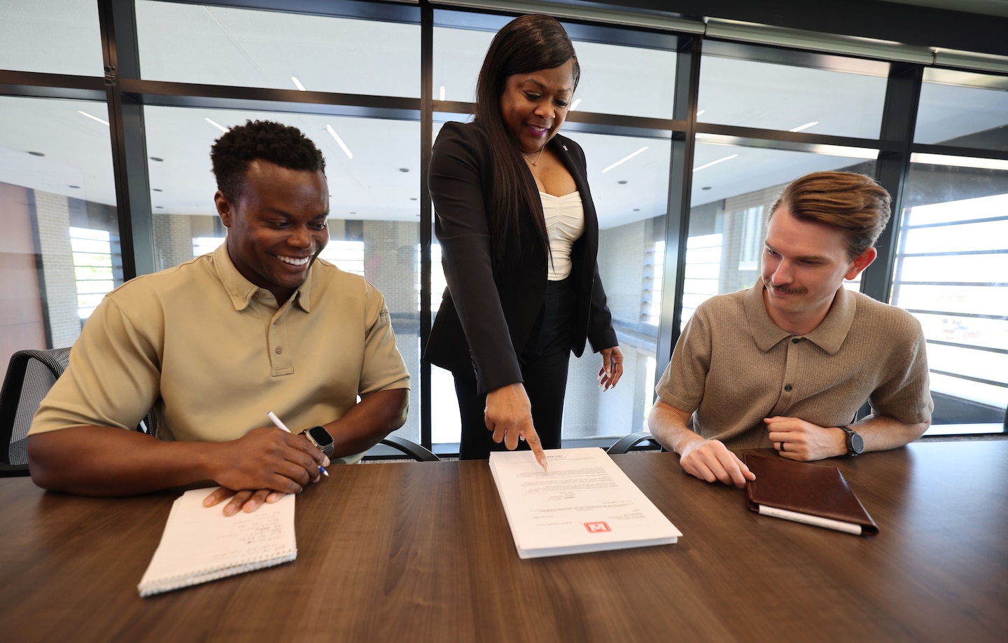 A woman standing point at a notepad talking to two men.