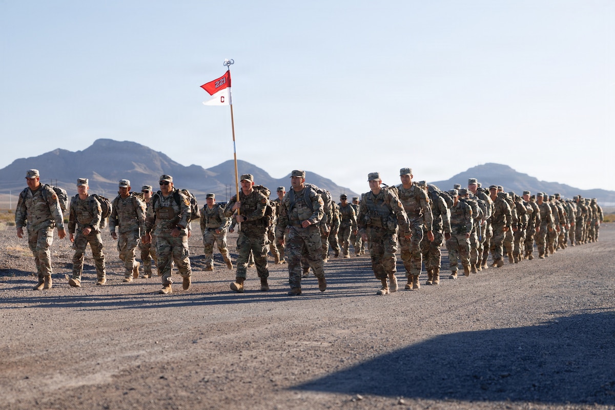 Dozens of people in camouflage military uniforms march in formation on a dirt road, with mountains in the background.
