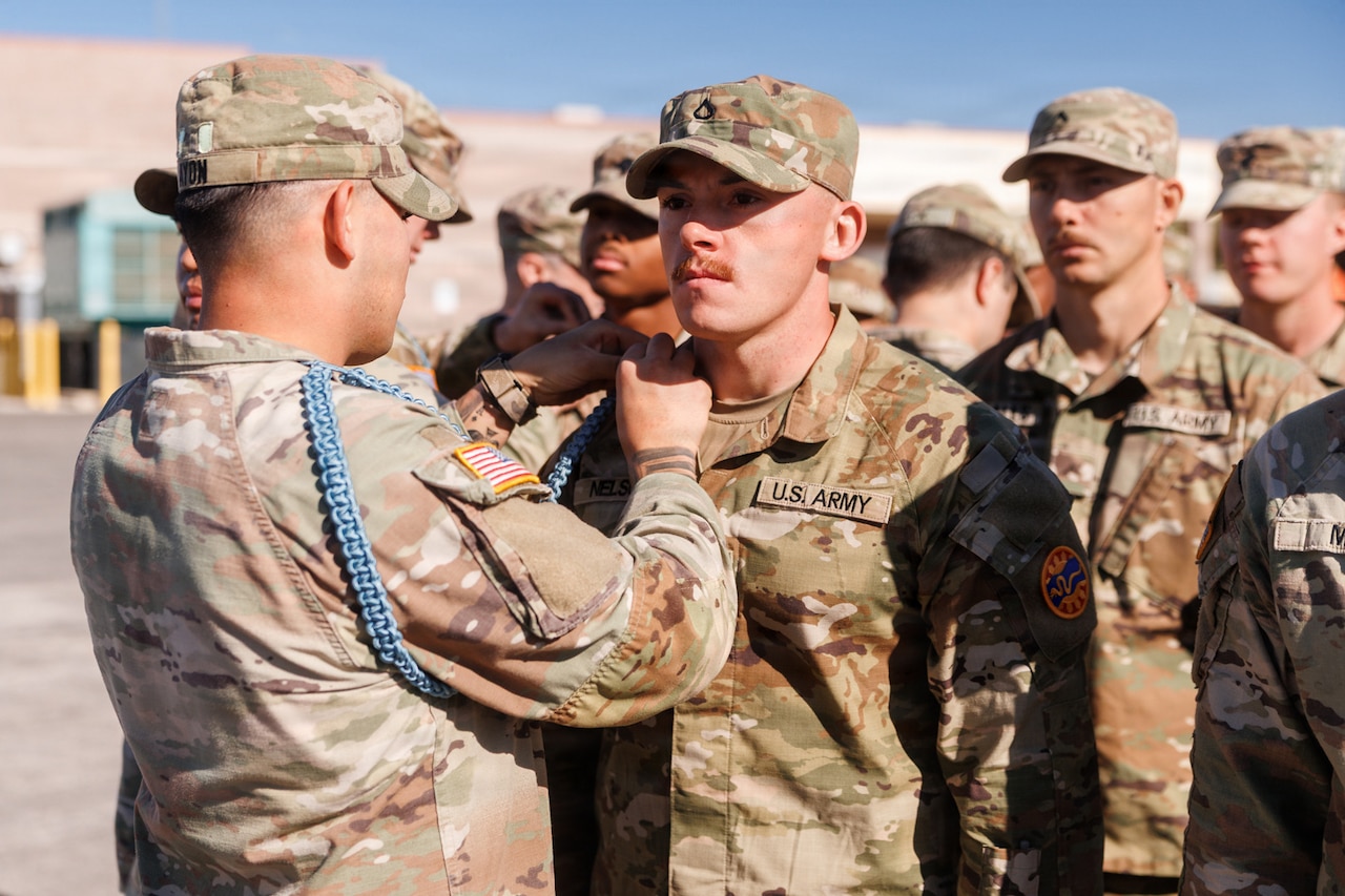 A man in a camouflage military uniform pins a rope onto the shoulder of another man in similar attire outside, while dozens of people, also in military uniforms, stand behind them.