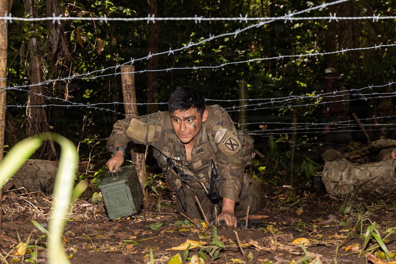 A man in a camouflage military uniform crawls in the jungle under barbed wire while holding an ammunition cannister.