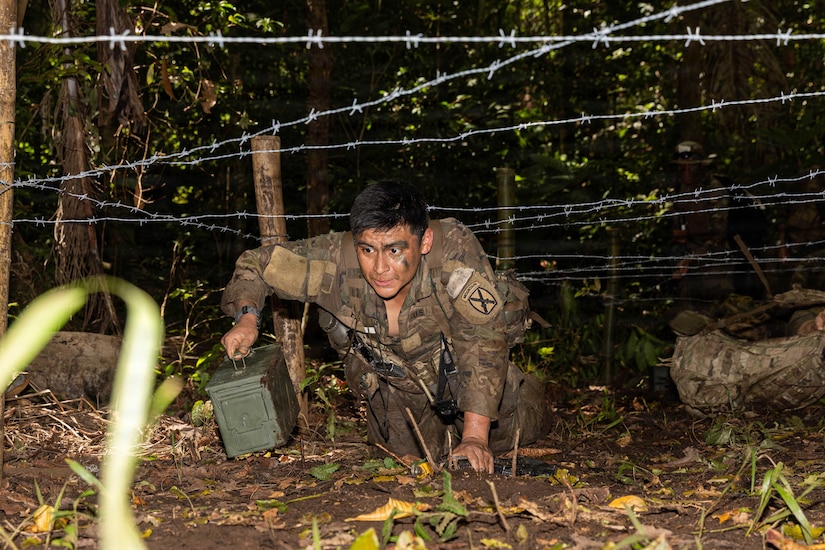 A man in a camouflage military uniform crawls in the jungle under barbed wire while holding an ammunition cannister.