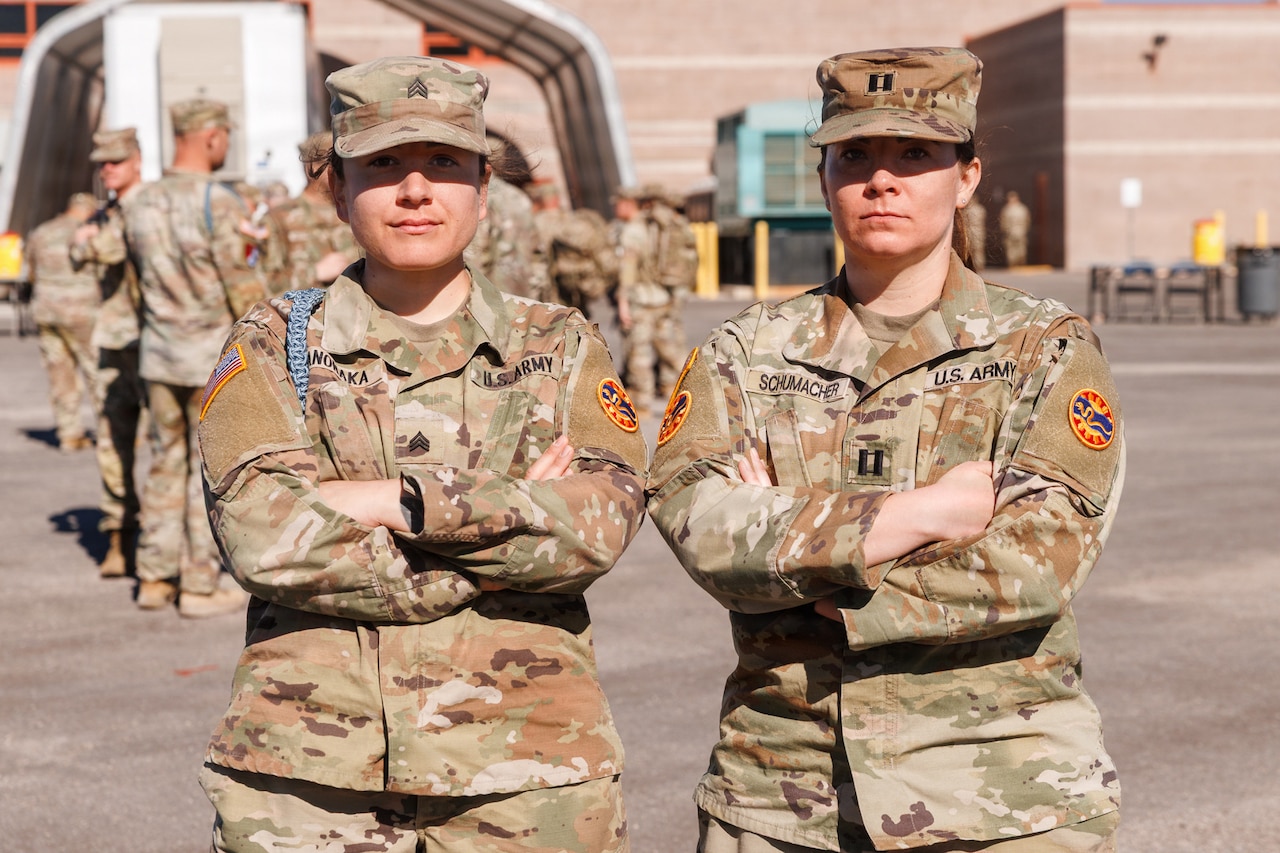 Two women in camouflage military uniforms pose for a photo with their arms crossed outside.