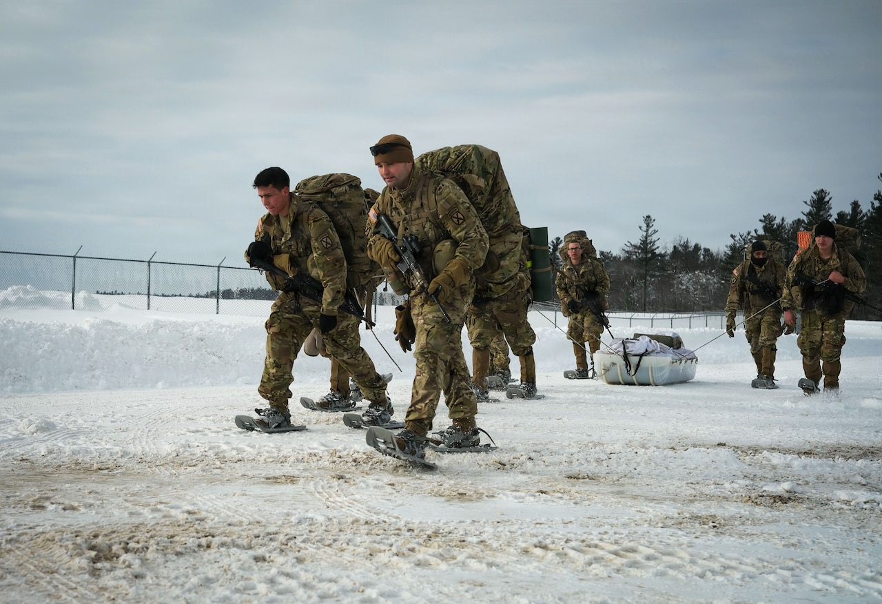 Six people in camouflage military uniforms and snowshoes walk on a snowy road while two other people in similar attire pull a sled filled with supplies.
