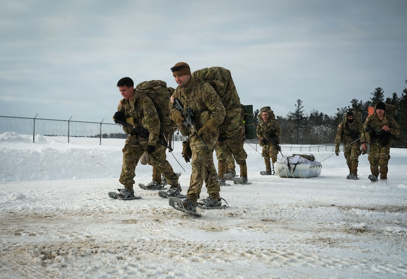 Six people in camouflage military uniforms and snowshoes walk on a snowy road while two other people in similar attire pull a sled filled with supplies.