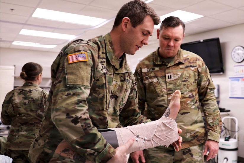 In a medical facility, a man in a camouflage military uniform checks on another person's foot that is wrapped in a splint, while another person in similar attire observes.