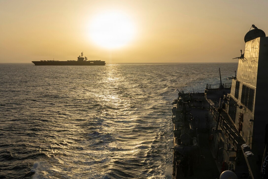 A ship, shown in silhouette, operates in the distance under a low sun against orangish sky, with part of another ship in the foreground.