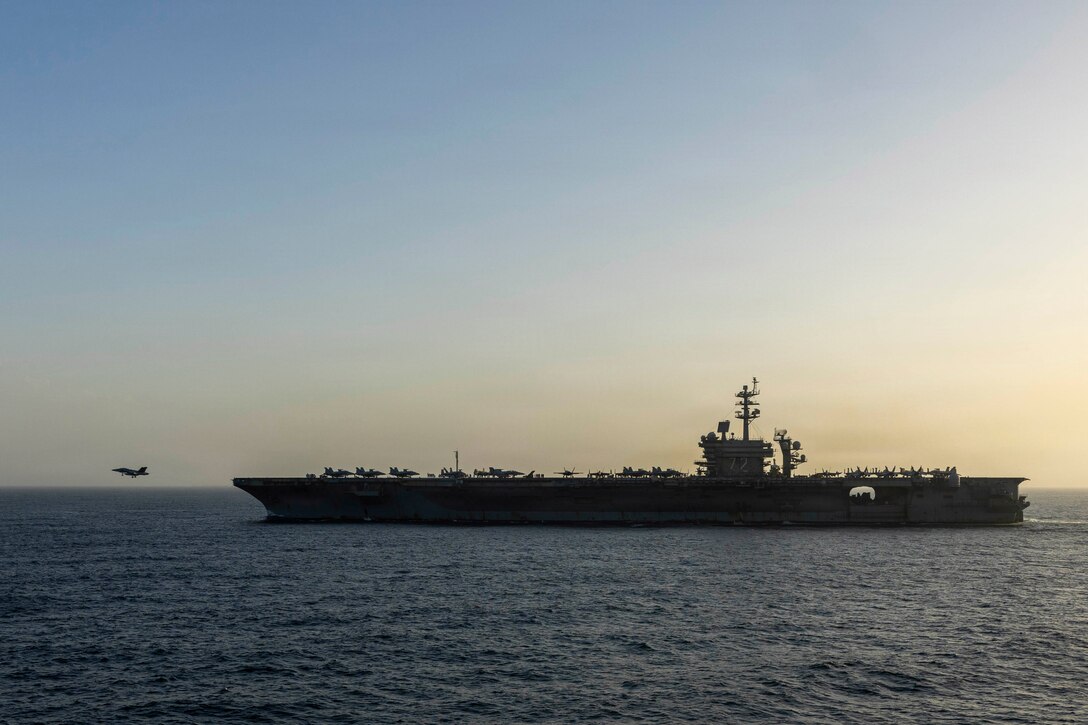 An aircraft flies in front of a ship in water, with orange and blue-tinged sky in the background.