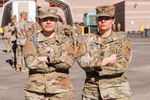 Two women in camouflage military uniforms pose for a photo with their arms crossed outside.