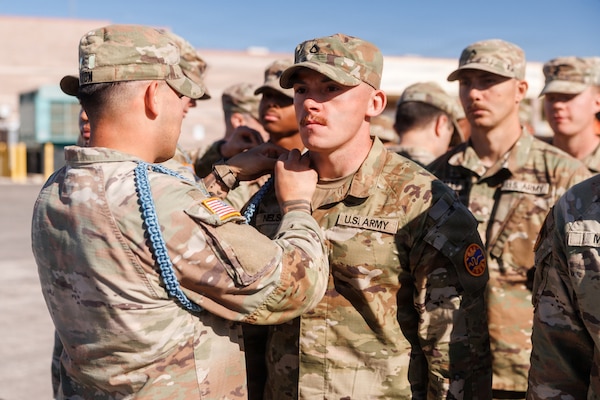 A man in a camouflage military uniform pins a rope onto the shoulder of another man in similar attire outside, while dozens of people, also in military uniforms, stand behind them.