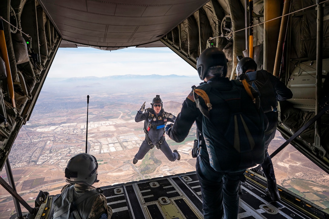 Three airmen watch a fellow airman descend over a city from the open doorway of an aircraft with mountains in the distance.