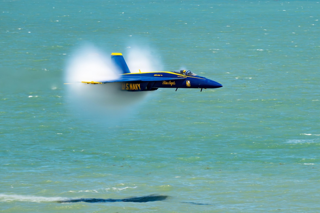 An aircraft flies over clear beach water, leaving behind a cloud of smoke during the day.