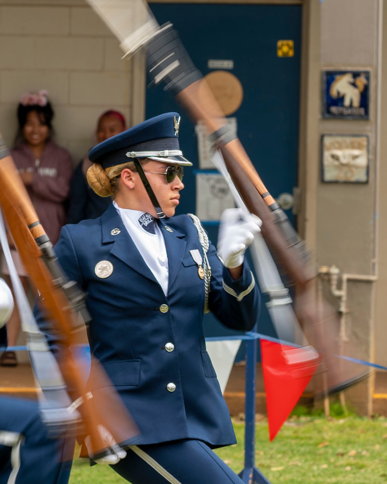 An Airman spins a rifle with students watching her in the background.