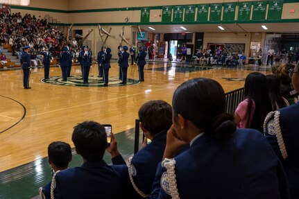 The drill team throws rifles in the air from the over-the-shoulder perspective of JROTC cadets wearing similar uniforms sitting in the bleachers.