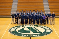 Approximately 25 individuals in blue Air Force uniforms pose for a group photo in a gym.