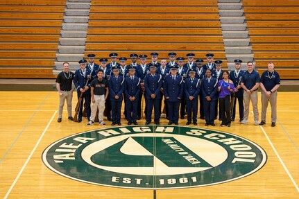 Approximately 25 individuals in blue Air Force uniforms pose for a group photo in a gym.