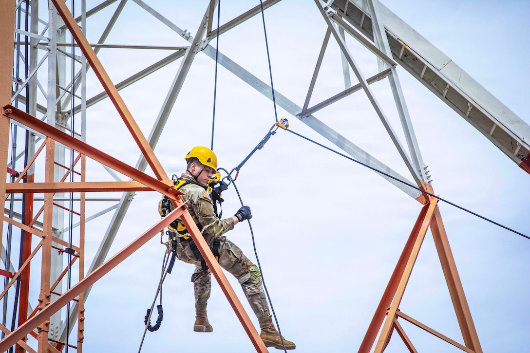A person in a camouflage uniform, yellow hard hat and harness hangs in the air from cables attached to a cellular tower and looks down.