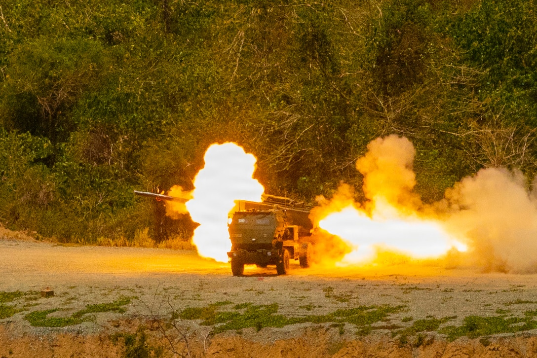 A rocket is fired from a vehicle parked on dirt, with smoke and fire seen on both sides of the vehicle and trees in the background.