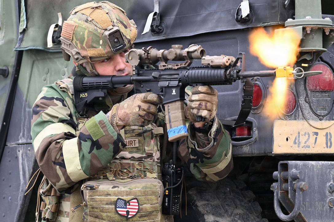 A person in a camouflage uniform fires an assault gun while kneeling by a tire on a military vehicle.