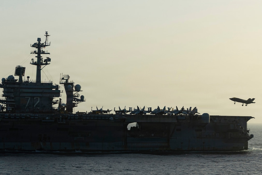 Aircraft sit on the deck of a military ship, with one jet in the air near an end of the ship, on a gray day.