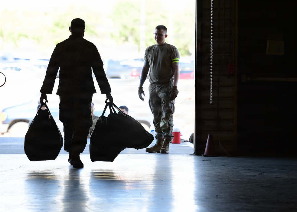 a silhouette photo depicts a male military member carrying two large bags at his sides out a warehouse doors to an evaluator standing just outside in the sun.