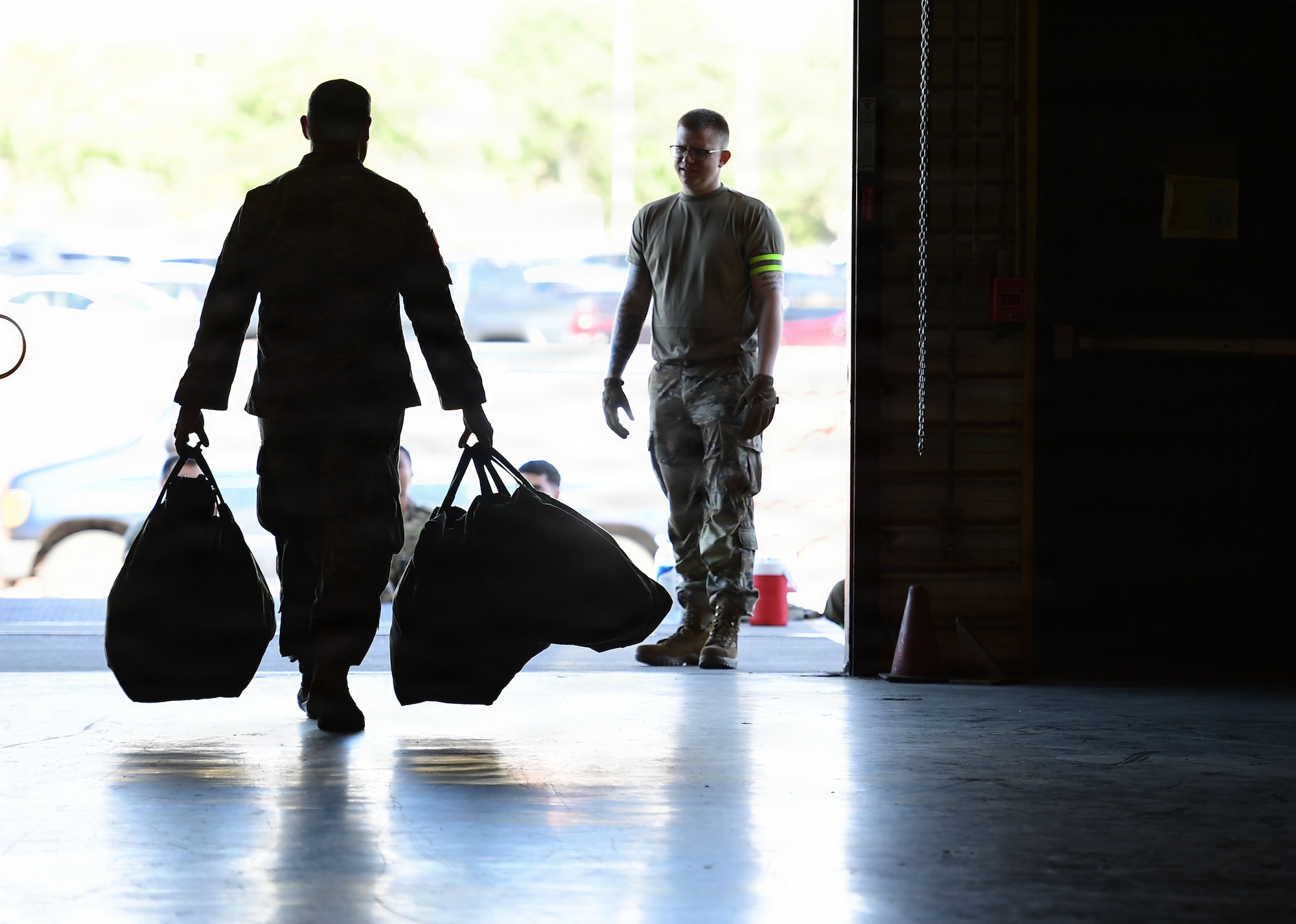 a silhouette photo depicts a male military member carrying two large bags at his sides out a warehouse doors to an evaluator standing just outside in the sun.