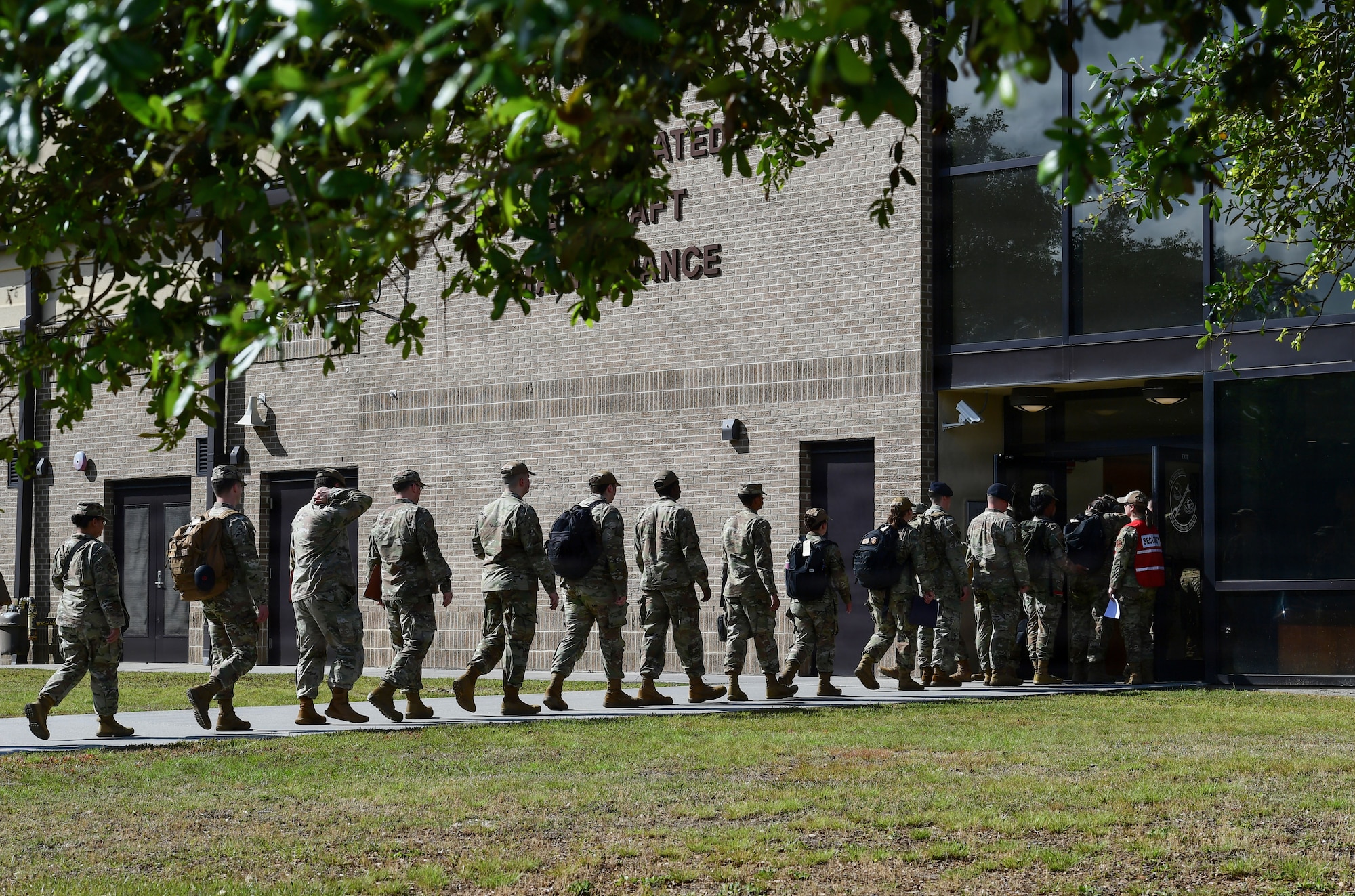 a wide shot depicts military personnel in uniform walking up a sidewalk into a brick building, with an overhead tree in the foreground