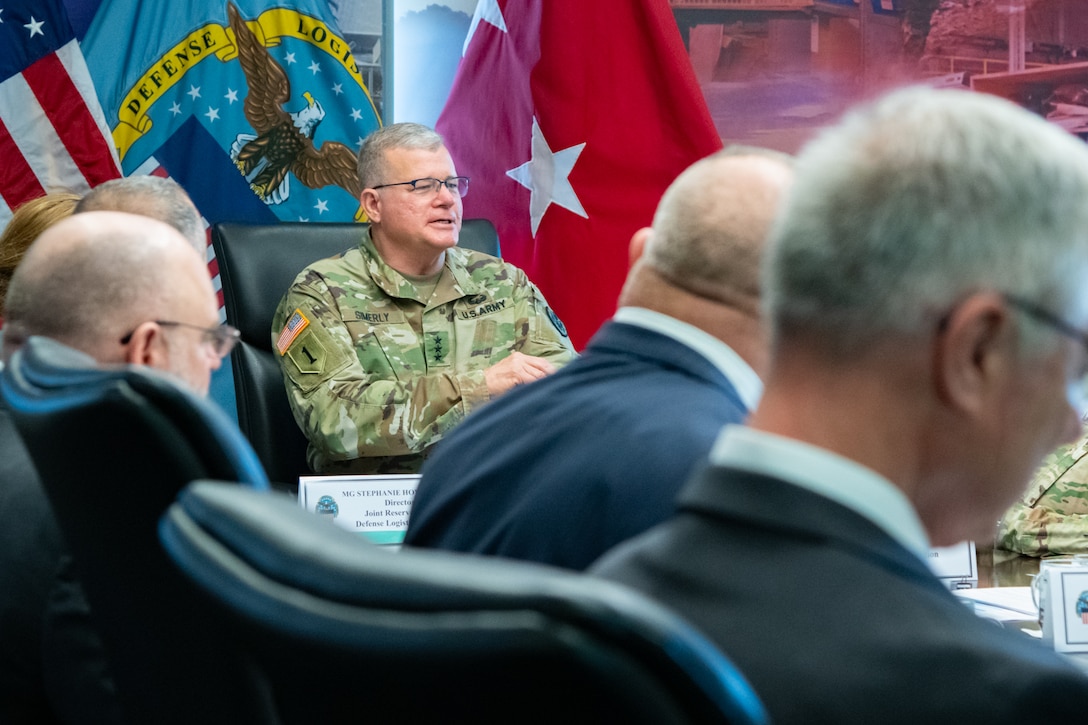 A man wearing a camouflage military uniform sits indoors at a conference room table with flags behind him speaking to a group of people also seated at the table.