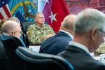 A man wearing a camouflage military uniform sits indoors at a conference room table with flags behind him speaking to a group of people also seated at the table.