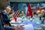 A man wearing a camouflage military uniform sits indoors at a conference room table with flags behind him speaking to a group of people also seated at the table.