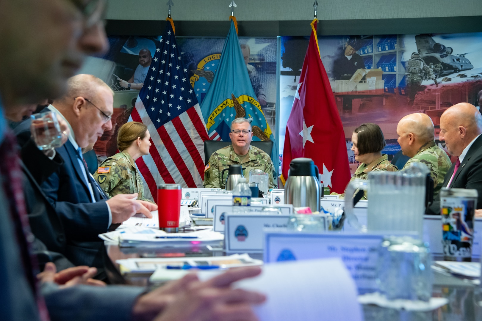 A man wearing a camouflage military uniform sits indoors at a conference room table with flags behind him speaking to a group of people also seated at the table.