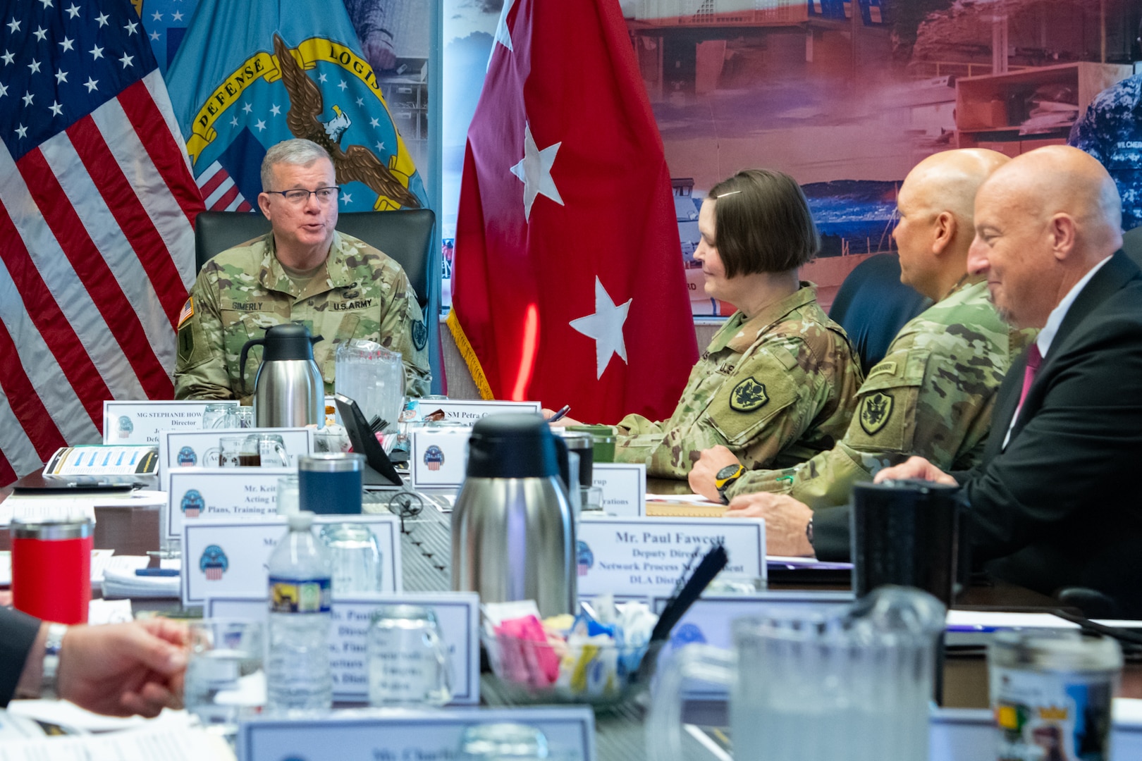 A man wearing a camouflage military uniform sits indoors at a conference room table with flags behind him speaking to a group of people.