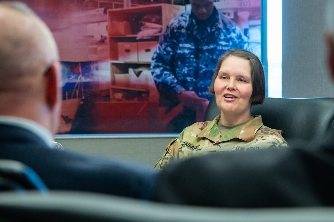 A woman wearing a camouflage military uniform sits indoors at a conference room table speaking to a group of people.