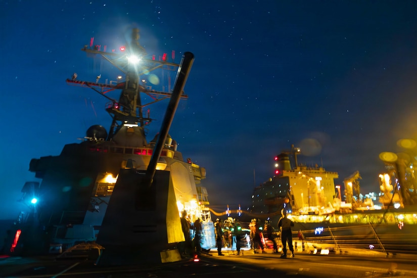 People in military uniforms stand aboard a large ship at night as another ship sails alongside them.