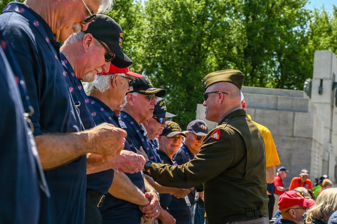 A soldier shakes hands with a veteran standing in line with fellow veterans in blue shirts on a sunny day.