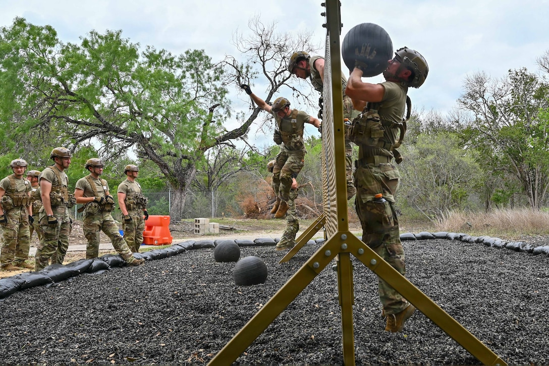 A candidate in tactical gear throws a weighted ball over an obstacle as fellow candidates climb over it while others watch from the left with a wooded area in the background.