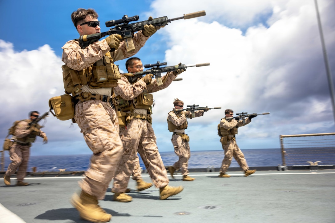 Marines aim rifles while walking in formation aboard a ship at sea during the day.