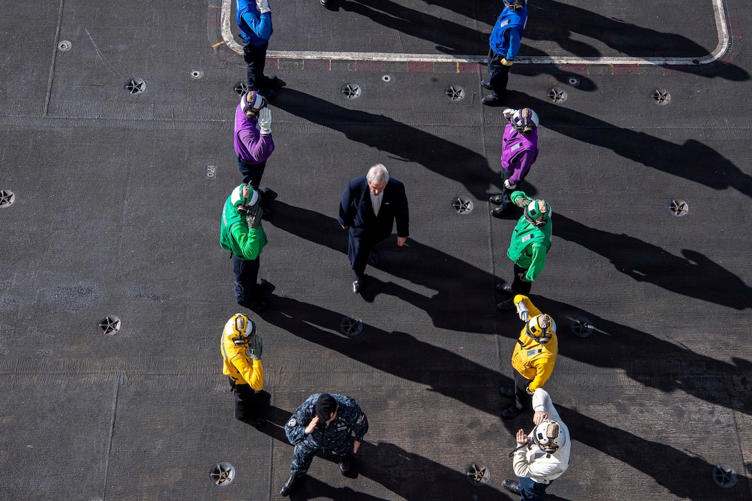 U.S. sailors in blue, purple, yellow and green vests stand in parallel lines on a ship at sea as Chilean officials walk in the center as seen from above.