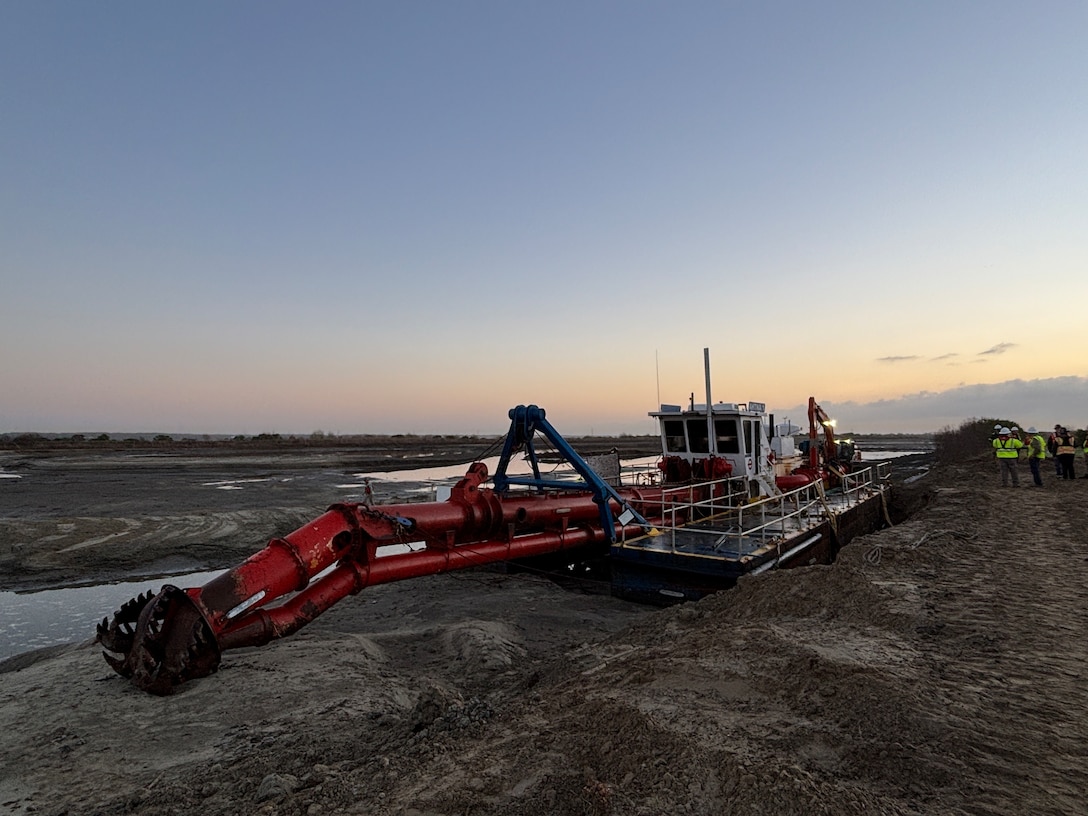 A barge with a dredge arms sitts on a sandy beach area, as a group stands in the distance beside it.