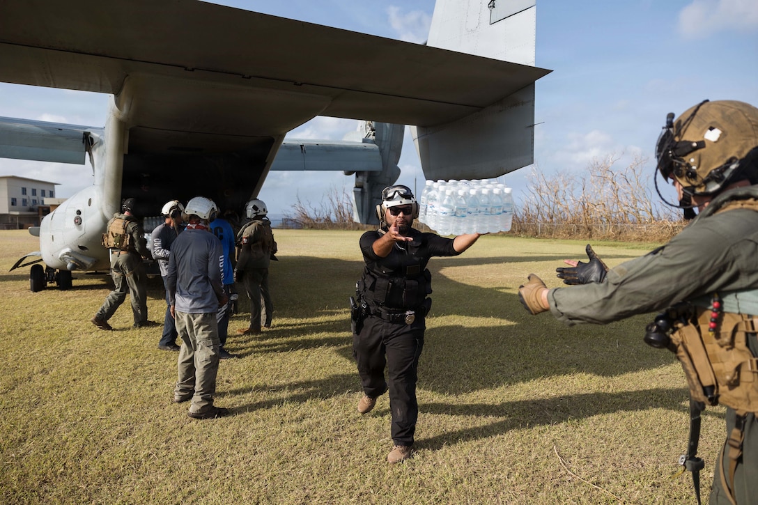 A person hands off water bottles to another person as a group of people stand behind a military aircraft parked on grass to unload more water bottles.