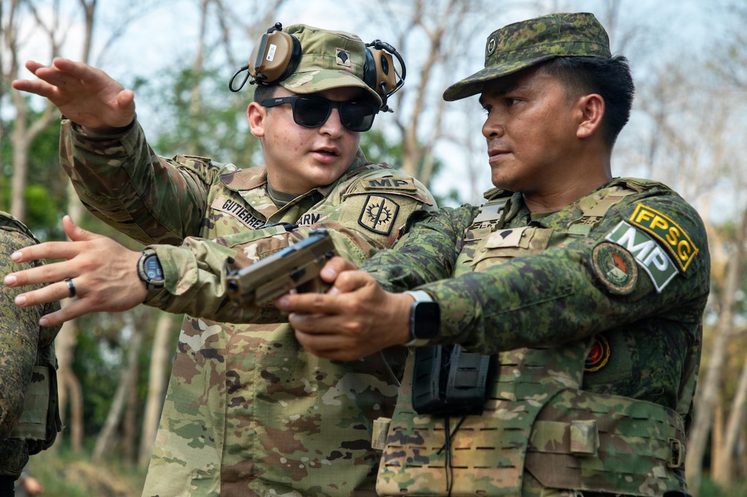 Two people in camouflage uniforms stand next to each other outside, with one person turning their head toward the other and making hand motions as the other person holds a gun.
