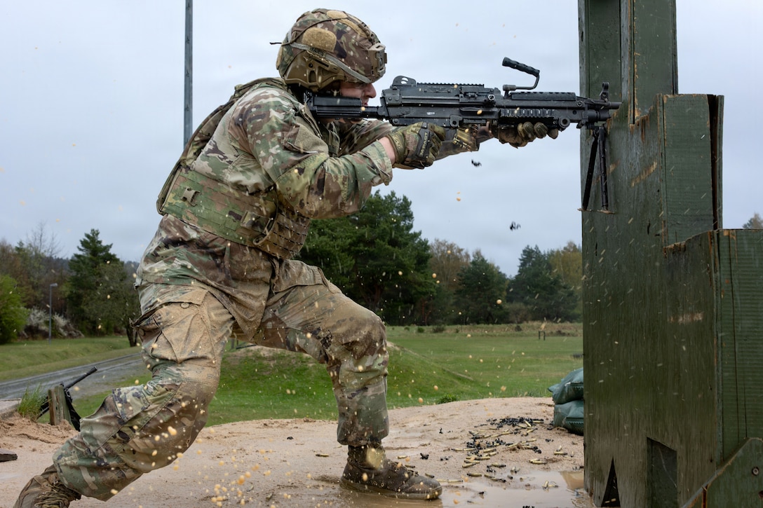 A person in a camouflage uniform stands in mud behind a wooden board and points a machine gun through an opening.