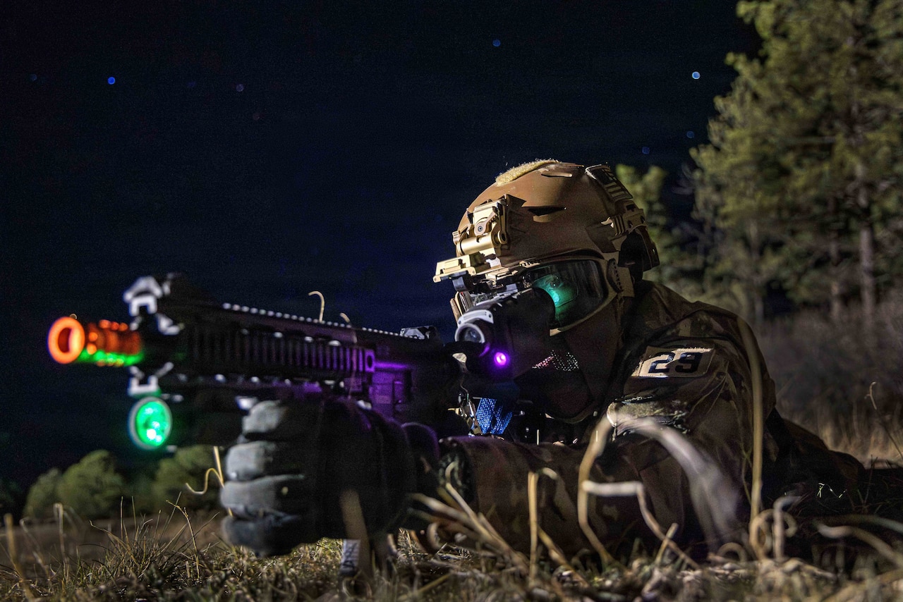 A person in a camouflage uniform and goggles lies on the grass in darkness with their torso lifted and points a machine gun.