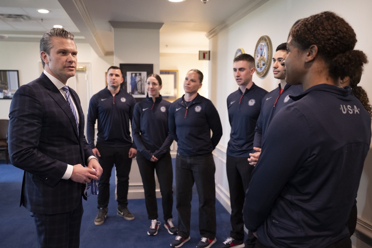 A man in business attire talks with a woman wearing a blue athletic jacket that has "USA" printed on the back. Others in similar attire stand nearby.