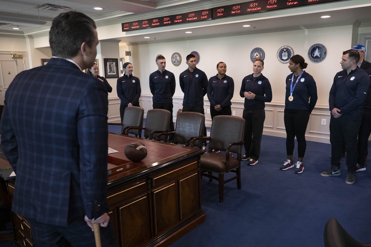 Nearly a dozen people in athletic attire stand in a conference room and speak with a man in business attire.